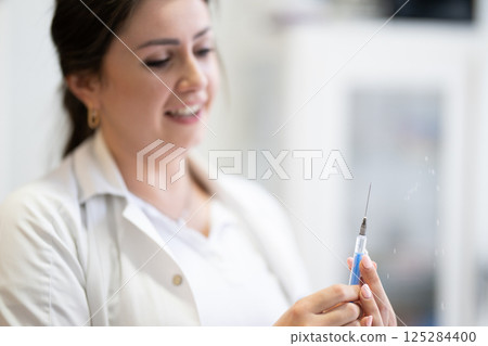 A focused nurse in a white lab coat carefully prepares a syringe for an injection in a professional healthcare environment. 125284400