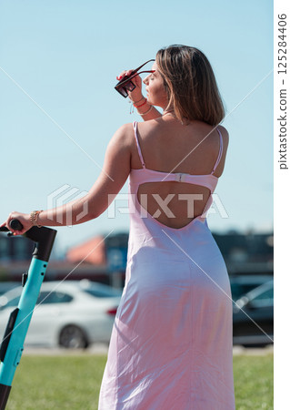 An elegant, stunning woman in a pink dress and sunglasses strikes a confident pose next to her electric scooter, radiating style and sophistication in the vibrant urban environment. 125284406