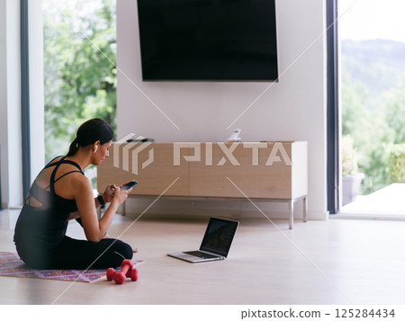A woman in sportswear is sitting in the living room and preparing for online training while using a laptop. A woman in sportswear is sitting in the living room and preparing for online training while using a laptop. 125284434