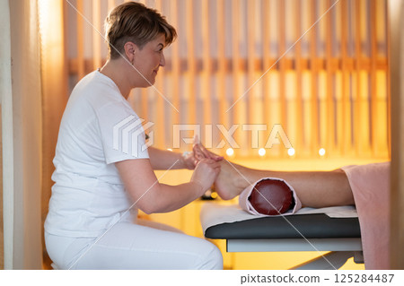 Foot Massage Therapy in a Relaxing Spa Setting. A professional massage therapist in a white uniform provides a soothing foot massage to a client in a calm and serene spa environment 125284487