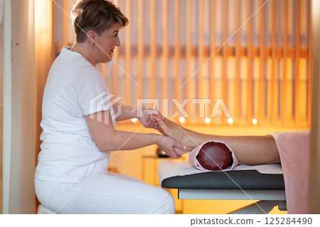 Foot Massage Therapy in a Relaxing Spa Setting. A professional massage therapist in a white uniform provides a soothing foot massage to a client in a calm and serene spa environment 125284490