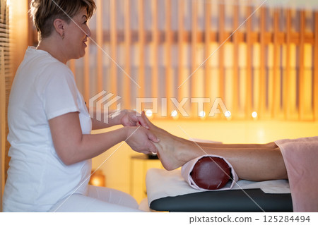 Foot Massage Therapy in a Relaxing Spa Setting. A professional massage therapist in a white uniform provides a soothing foot massage to a client in a calm and serene spa environment Foot Massage Therapy in a Relaxing Spa Setting. A professional massage therapist in a white uniform provides a soothing foot massage to a client in a calm and serene spa environment 125284494
