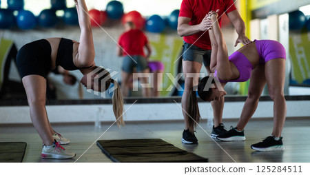 A trainer guides two women through stretching exercises in the gym, promoting flexibility, wellness, and a healthy lifestyle. 125284511