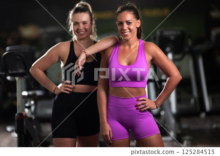 Two fitness women resting and recovering in the gym after an intense workout session, showcasing strength, endurance, and dedication to their fitness journey. Two fitness women resting and recovering in the gym after an intense workout session, showcasing strength, endurance, and dedication to their fitness journey. 125284515