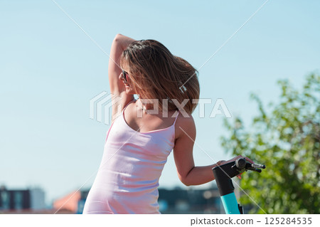 An elegant, stunning woman in a pink dress and sunglasses strikes a confident pose next to her electric scooter, radiating style and sophistication in the vibrant urban environment. 125284535