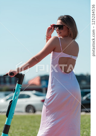 An elegant, stunning woman in a pink dress and sunglasses strikes a confident pose next to her electric scooter, radiating style and sophistication in the vibrant urban environment. 125284536