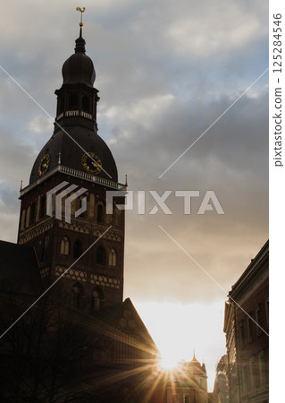 Historic Church Tower with Sunset in the Background.A striking view of a historic church tower silhouetted against a dramatic sky at sunset. The golden sunlight peeks through surrounding buildings 125284546