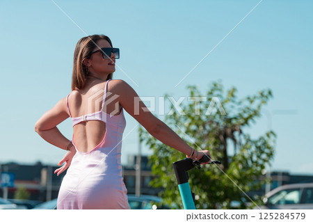 An elegant, stunning woman in a pink dress and sunglasses strikes a confident pose next to her electric scooter, radiating style and sophistication in the vibrant urban environment. An elegant, stunning woman in a pink dress and sunglasses strikes a confident pose next to her electric scooter, radiating style and sophistication in the vibrant urban environment. 125284579