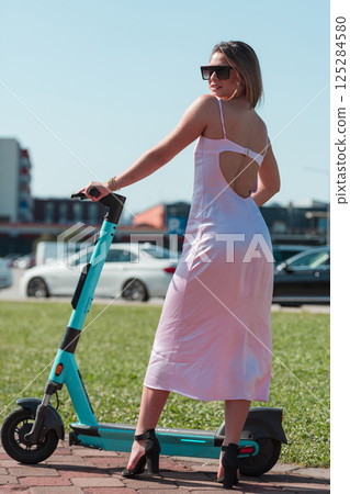 An elegant, stunning woman in a pink dress and sunglasses strikes a confident pose next to her electric scooter, radiating style and sophistication in the vibrant urban environment. 125284580