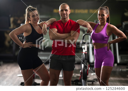 A determined fitness trainer standing confidently in front of his two female clients, guiding them through their workout in the background. A determined fitness trainer standing confidently in front of his two female clients, guiding them through their workout in the background. 125284600