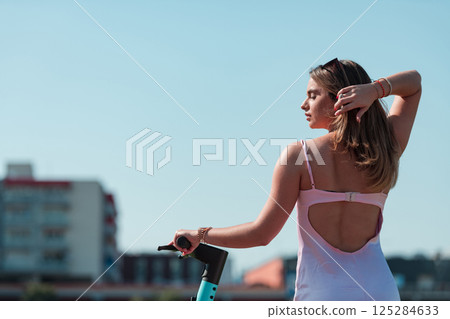 An elegant, stunning woman in a pink dress and sunglasses strikes a confident pose next to her electric scooter, radiating style and sophistication in the vibrant urban environment. 125284633