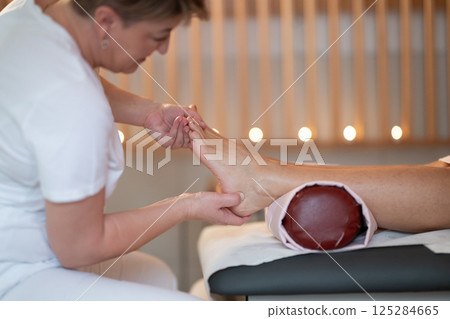 Foot Massage Therapy in a Relaxing Spa Setting. A professional massage therapist in a white uniform provides a soothing foot massage to a client in a calm and serene spa environment 125284665