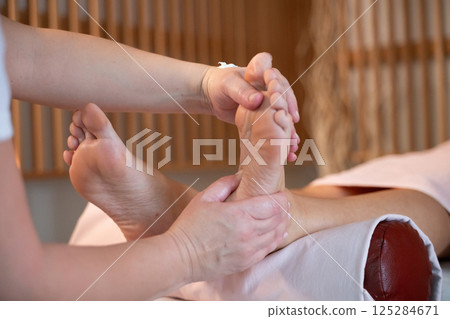 Foot Massage Therapy in a Relaxing Spa Setting. A professional massage therapist in a white uniform provides a soothing foot massage to a client in a calm and serene spa environment Foot Massage Therapy in a Relaxing Spa Setting. A professional massage therapist in a white uniform provides a soothing foot massage to a client in a calm and serene spa environment 125284671