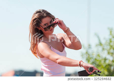 An elegant, stunning woman in a pink dress and sunglasses strikes a confident pose next to her electric scooter, radiating style and sophistication in the vibrant urban environment. 125284682