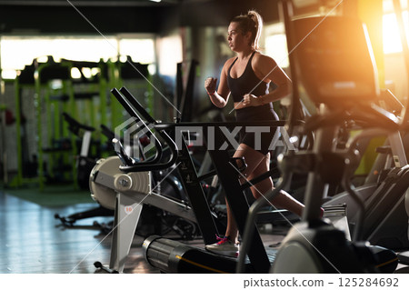 Woman Running on Treadmill in Gym.A fit woman wearing a purple sports outfit is running on a treadmill in a gym. She has a focused yet relaxed expression, with gym equipment and a warm light in the 125284692