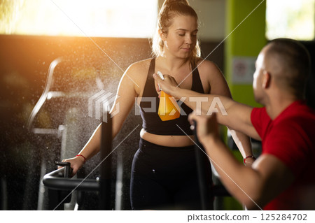 A man carefully applies a sweat simulating spray to a woman as he prepares her for a fitness photoshoot, ensuring every detail is perfect for the session. 125284702