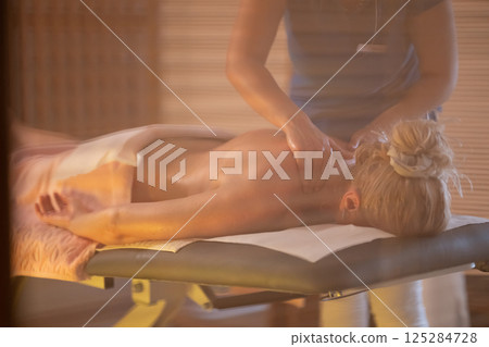 Relaxing Back Massage in a Spa Setting. A professional massage therapist in a blue uniform provides a relaxing back massage to a client lying on a massage table. 125284728