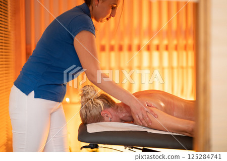 Relaxing Back Massage in a Spa Setting. A professional massage therapist in a blue uniform provides a relaxing back massage to a client lying on a massage table. 125284741