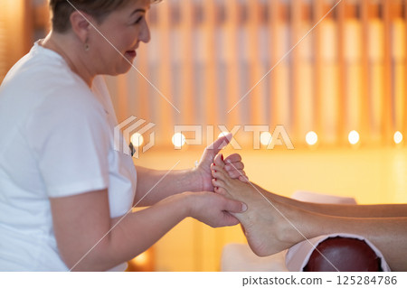 Foot Massage Therapy in a Relaxing Spa Setting. A professional massage therapist in a white uniform provides a soothing foot massage to a client in a calm and serene spa environment 125284786