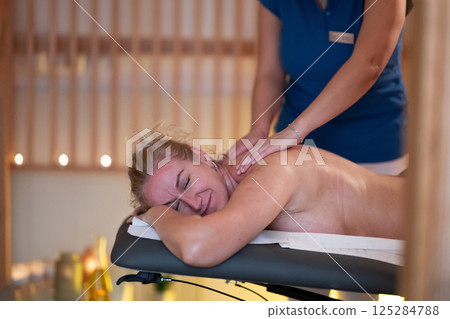 Relaxing Back Massage in a Spa Setting. A professional massage therapist in a blue uniform provides a relaxing back massage to a client lying on a massage table. Relaxing Back Massage in a Spa Setting. A professional massage therapist in a blue uniform provides a relaxing back massage to a client lying on a massage table. 125284788