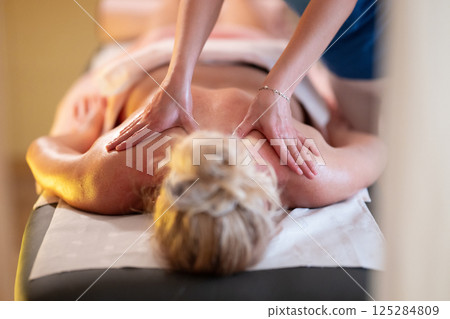 Relaxing Back Massage in a Spa Setting. A professional massage therapist in a blue uniform provides a relaxing back massage to a client lying on a massage table. Relaxing Back Massage in a Spa Setting. A professional massage therapist in a blue uniform provides a relaxing back massage to a client lying on a massage table. 125284809