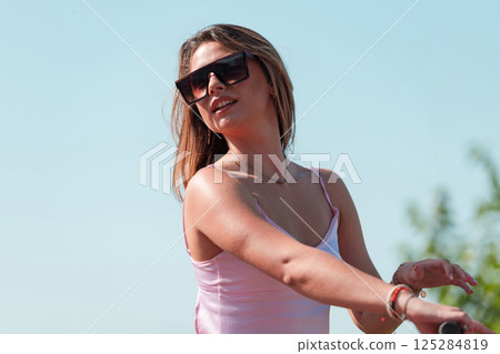 An elegant, stunning woman in a pink dress and sunglasses strikes a confident pose next to her electric scooter, radiating style and sophistication in the vibrant urban environment. An elegant, stunning woman in a pink dress and sunglasses strikes a confident pose next to her electric scooter, radiating style and sophistication in the vibrant urban environment. 125284819