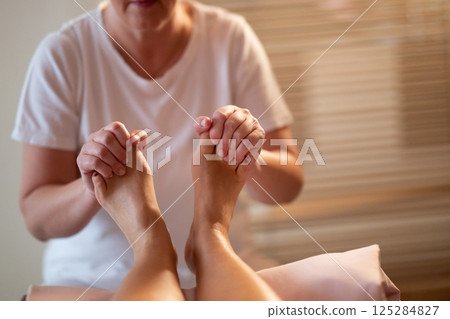 Foot Massage Therapy in a Relaxing Spa Setting. A professional massage therapist in a white uniform provides a soothing foot massage to a client in a calm and serene spa environment 125284827