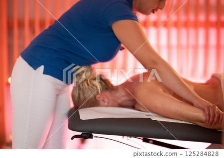 Relaxing Back Massage in a Spa Setting. A professional massage therapist in a blue uniform provides a relaxing back massage to a client lying on a massage table. 125284828