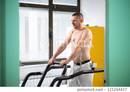 A middle-aged man is undergoing a medical stress test on a treadmill, connected to ECG electrodes for heart monitoring in a clinical environment A middle-aged man is undergoing a medical stress test on a treadmill, connected to ECG electrodes for heart monitoring in a clinical environment 125284837