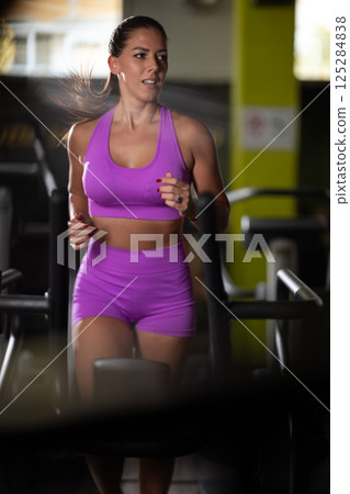 Woman Running on Treadmill in Gym.A fit woman wearing a purple sports outfit is running on a treadmill in a gym. She has a focused yet relaxed expression, with gym equipment and a warm light in the Woman Running on Treadmill in Gym.A fit woman wearing a purple sports outfit is running on a treadmill in a gym. She has a focused yet relaxed expression, with gym equipment and a warm light in the 125284838