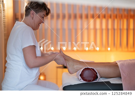 Foot Massage Therapy in a Relaxing Spa Setting. A professional massage therapist in a white uniform provides a soothing foot massage to a client in a calm and serene spa environment Foot Massage Therapy in a Relaxing Spa Setting. A professional massage therapist in a white uniform provides a soothing foot massage to a client in a calm and serene spa environment 125284849