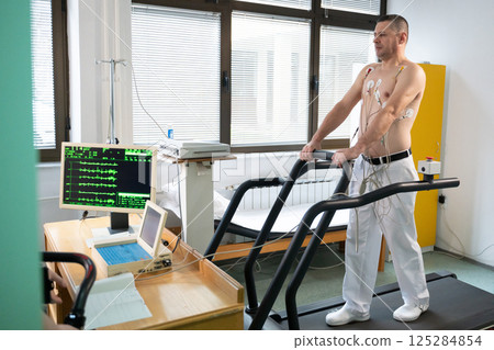 A middle-aged man is undergoing a medical stress test on a treadmill, connected to ECG electrodes for heart monitoring in a clinical environment A middle-aged man is undergoing a medical stress test on a treadmill, connected to ECG electrodes for heart monitoring in a clinical environment 125284854