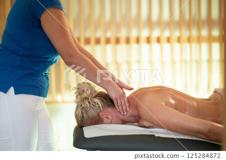 Relaxing Back Massage in a Spa Setting. A professional massage therapist in a blue uniform provides a relaxing back massage to a client lying on a massage table. 125284872