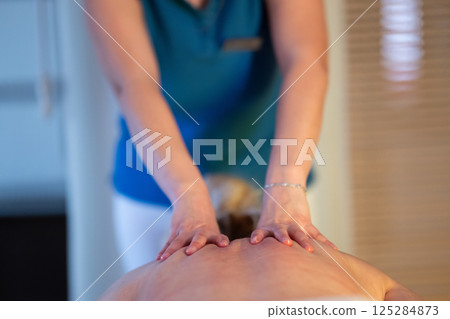 Relaxing Back Massage in a Spa Setting. A professional massage therapist in a blue uniform provides a relaxing back massage to a client lying on a massage table. Relaxing Back Massage in a Spa Setting. A professional massage therapist in a blue uniform provides a relaxing back massage to a client lying on a massage table. 125284873