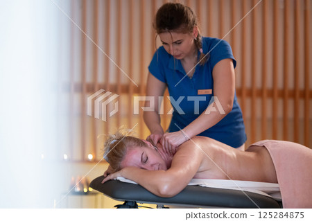 Relaxing Back Massage in a Spa Setting. A professional massage therapist in a blue uniform provides a relaxing back massage to a client lying on a massage table. Relaxing Back Massage in a Spa Setting. A professional massage therapist in a blue uniform provides a relaxing back massage to a client lying on a massage table. 125284875