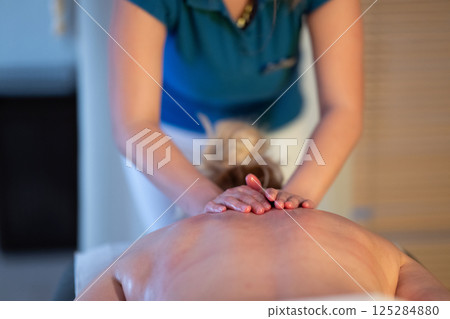Relaxing Back Massage in a Spa Setting. A professional massage therapist in a blue uniform provides a relaxing back massage to a client lying on a massage table. 125284880