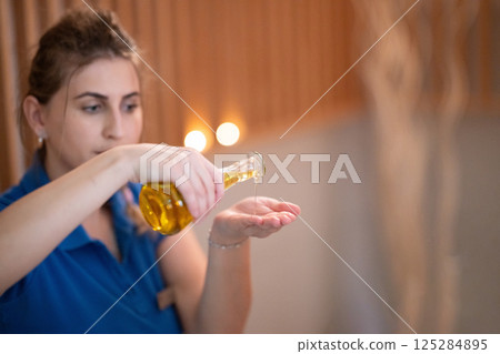 A professional massage therapist pours aromatic oil into their hands, preparing for a relaxing and rejuvenating massage session in a serene spa setting. 125284895