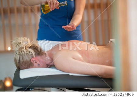 A professional massage therapist pours aromatic oil into their hands, preparing for a relaxing and rejuvenating massage session in a serene spa setting. 125284909