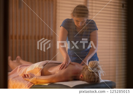 Relaxing Back Massage in a Spa Setting. A professional massage therapist in a blue uniform provides a relaxing back massage to a client lying on a massage table. 125285015