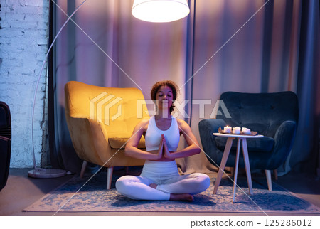 An attractive young African in a white fitness suit sits in a lotus position and relaxes. A cute black girl is meditating in the living room. A relaxed woman does evening yoga at home. An attractive young African in a white fitness suit sits in a lotus position and relaxes. A cute black girl is meditating in the living room. A relaxed woman does evening yoga at home. 125286012