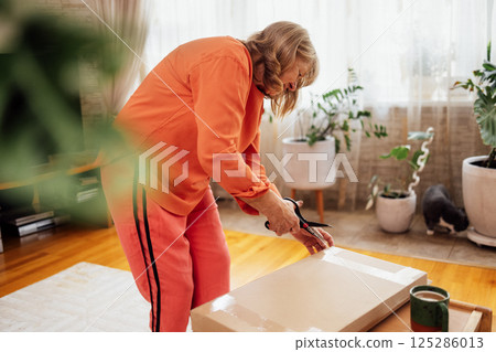 Elderly woman in orange casual clothes unpacks package. Smiling middle-aged lady opens cardboard box in living room. Online shopping and delivery of goods by courier at home. Elderly woman in orange casual clothes unpacks package. Smiling middle-aged lady opens cardboard box in living room. Online shopping and delivery of goods by courier at home. 125286013