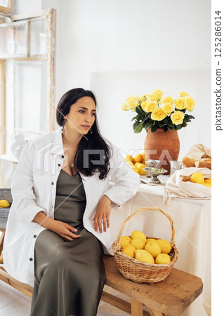 Young woman smiles and sits at set table with lemons. Clay pitcher with yellow roses 125286014