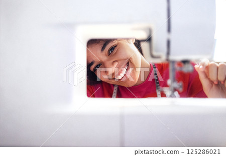 Close-up of a laughing African woman peeking out from behind a sewing machine. A smiling black girl in a sweater with a measuring tape and needle. 125286021