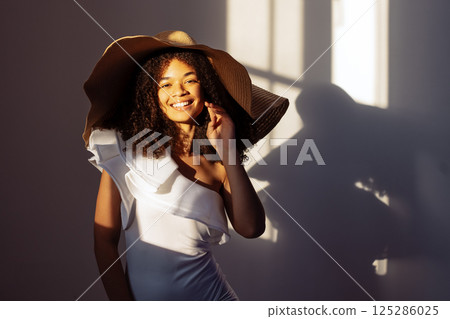Close-up portrait of a young attractive smiling African woman in an elegant straw hat with a wide brim. A charming black girl in a white fancy dress is laughing and posing. Close-up portrait of a young attractive smiling African woman in an elegant straw hat with a wide brim. A charming black girl in a white fancy dress is laughing and posing. 125286025