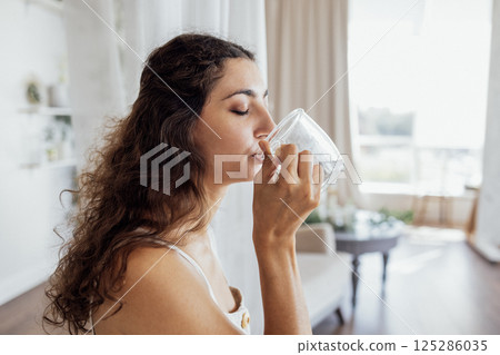 A charming curly-haired brunette in a linen sundress holds a transparent cup and drinks water 125286035