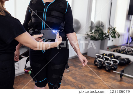 Close up of trainers hands help to put on ems suit to female plump client in fitness club. Close up of trainers hands help to put on ems suit to female plump client in fitness club. 125286079