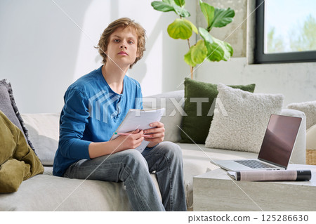 Teenager guy student studying sitting at home on sofa using laptop textbook notebook Teenager guy student studying sitting at home on sofa using laptop textbook notebook 125286630