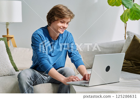 Handsome smiling teenage boy sitting at home on the couch typing on laptop Handsome smiling teenage boy sitting at home on the couch typing on laptop 125286631