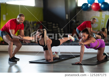 A trainer guides two women through stretching exercises in the gym, promoting flexibility, wellness, and a healthy lifestyle. 125287173