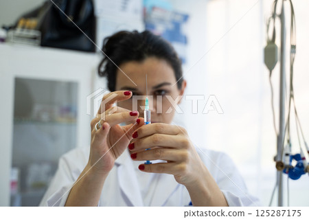 A focused nurse in a white lab coat carefully prepares a syringe for an injection in a professional healthcare environment. 125287175